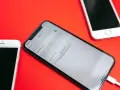 Tre iPhone su sfondo rosso. Uno mostra la schermata di backup iCloud in corso, con un cavo collegato.