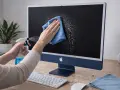 Mani che spruzzano liquido per pulire schermo pc con un panno azzurro su un iMac blu.