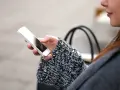 Mano che tiene uno smartphone, con un maglione a trecce e una borsa sullo sfondo. La persona sta utilizzando dati per navigare.
