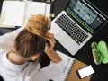 Donna frustrata con la testa tra le mani, laptop aperto, agenda e occhiali. La connessione lenta sta rendendo il lavoro impossibile.
