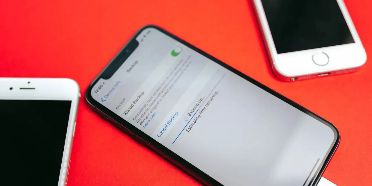 Tre iPhone su sfondo rosso. Uno mostra la schermata di backup iCloud in corso, con un cavo collegato.