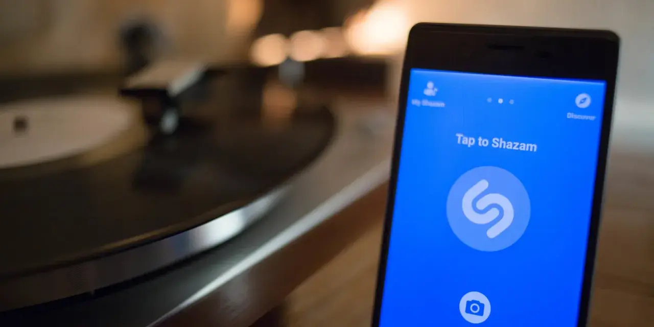 Smartphone con app Shazam aperta, pronto a identificare un brano. Sullo sfondo, un giradischi vintage.