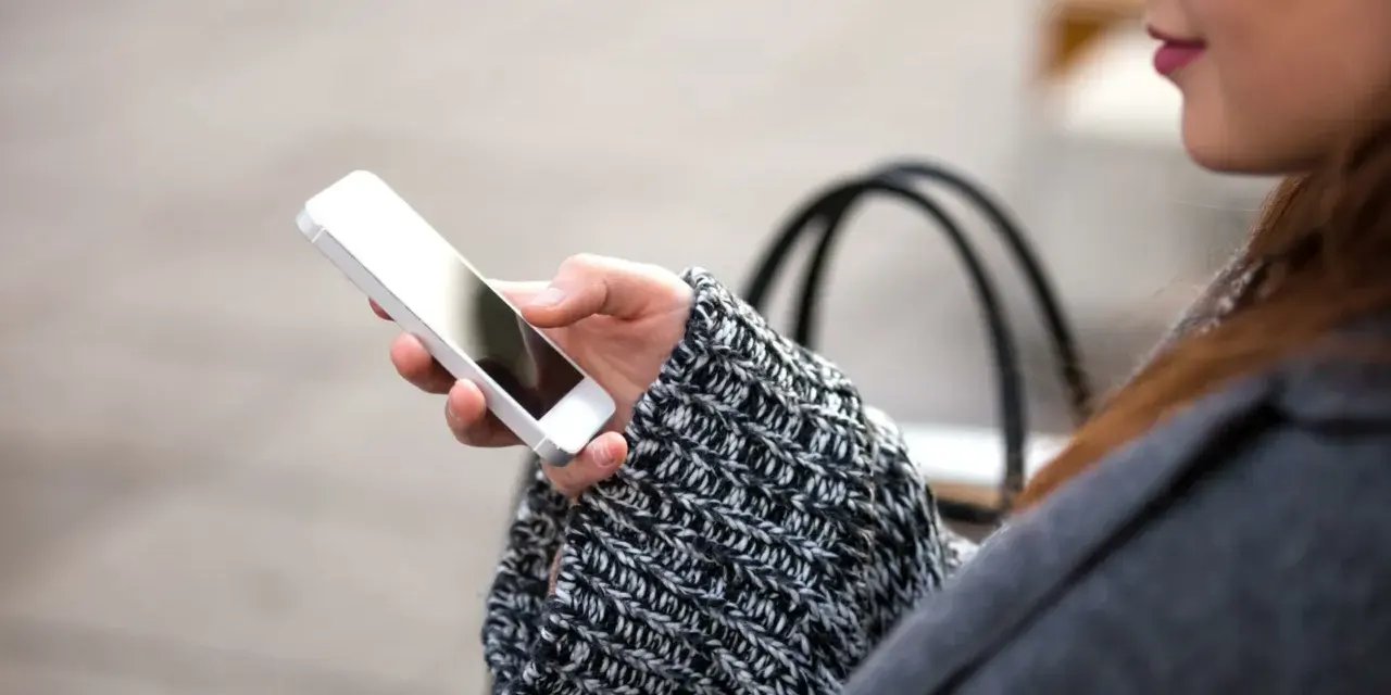 Mano che tiene uno smartphone, con un maglione a trecce e una borsa sullo sfondo. La persona sta utilizzando dati per navigare.