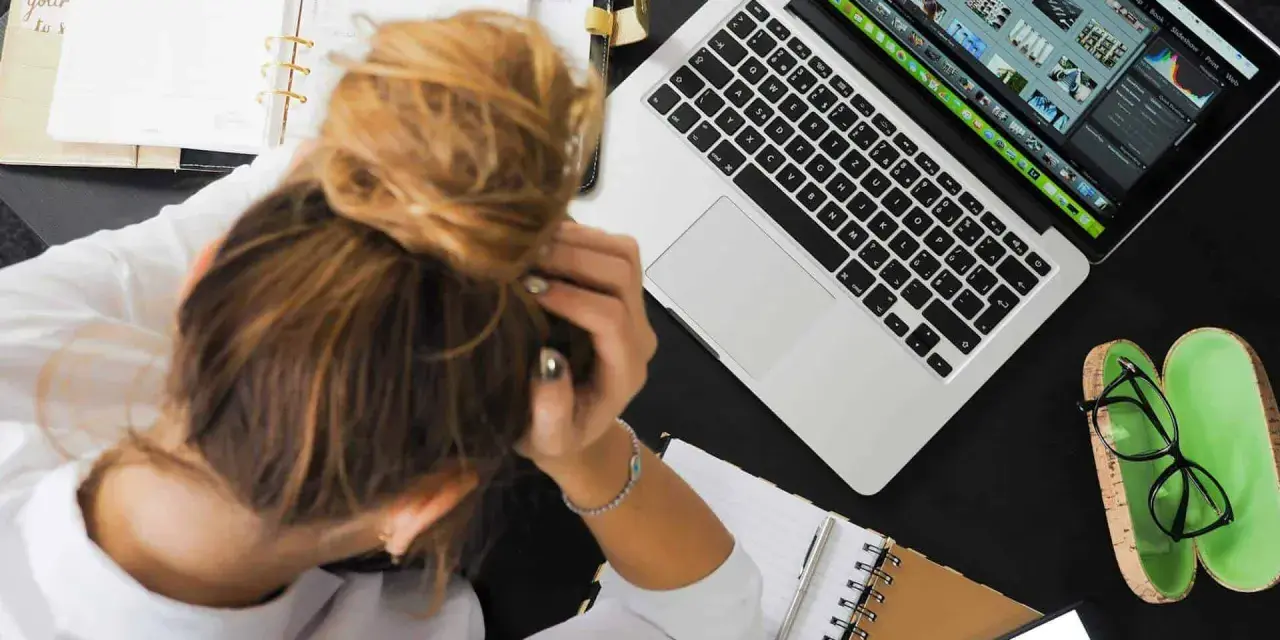 Donna frustrata con la testa tra le mani, laptop aperto, agenda e occhiali. La connessione lenta sta rendendo il lavoro impossibile.