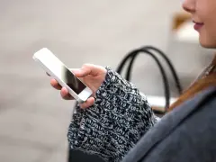 Mano che tiene uno smartphone, con un maglione a trecce e una borsa sullo sfondo. La persona sta utilizzando dati per navigare.