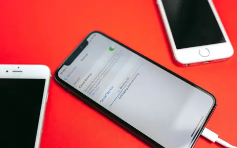 Tre iPhone su sfondo rosso. Uno mostra la schermata di backup iCloud in corso, con un cavo collegato.