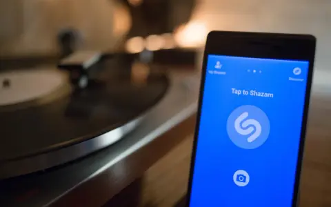 Smartphone con app Shazam aperta, pronto a identificare un brano. Sullo sfondo, un giradischi vintage.