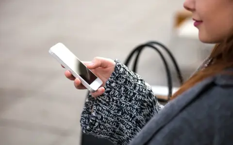 Mano che tiene uno smartphone, con un maglione a trecce e una borsa sullo sfondo. La persona sta utilizzando dati per navigare.