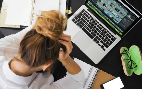 Donna frustrata con la testa tra le mani, laptop aperto, agenda e occhiali. La connessione lenta sta rendendo il lavoro impossibile.