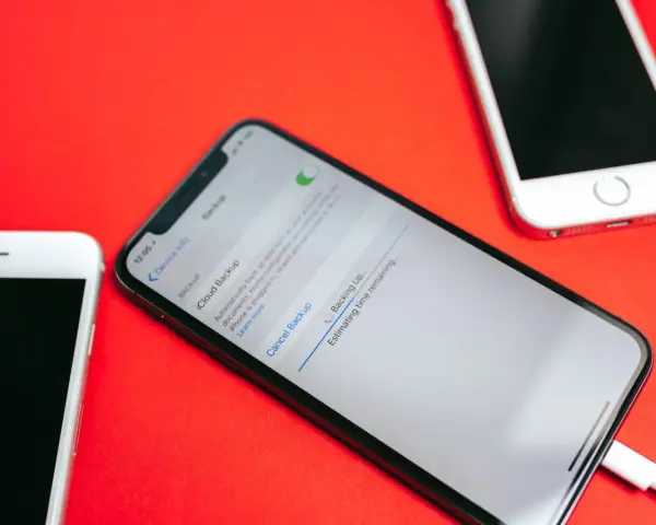 Tre iPhone su sfondo rosso. Uno mostra la schermata di backup iCloud in corso, con un cavo collegato.