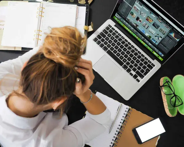 Donna frustrata con la testa tra le mani, laptop aperto, agenda e occhiali. La connessione lenta sta rendendo il lavoro impossibile.