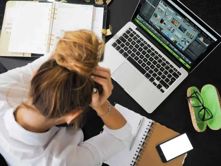 Donna frustrata con la testa tra le mani, laptop aperto, agenda e occhiali. La connessione lenta sta rendendo il lavoro impossibile.