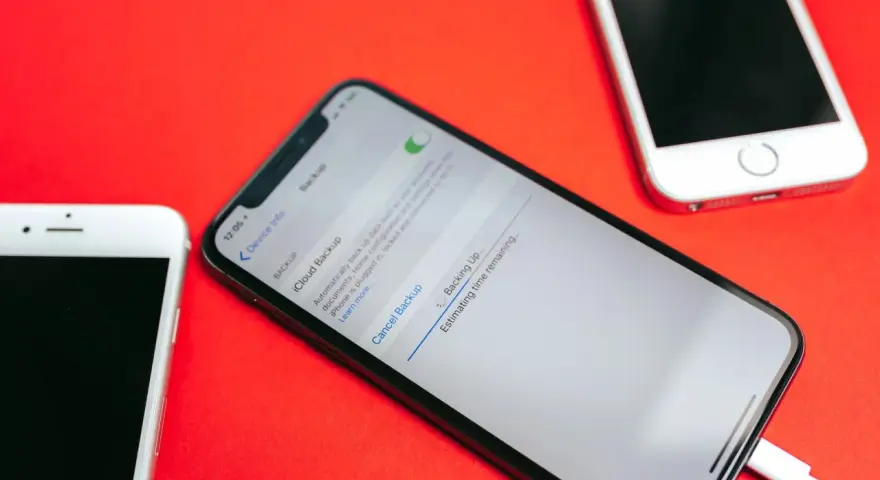 Tre iPhone su sfondo rosso. Uno mostra la schermata di backup iCloud in corso, con un cavo collegato.