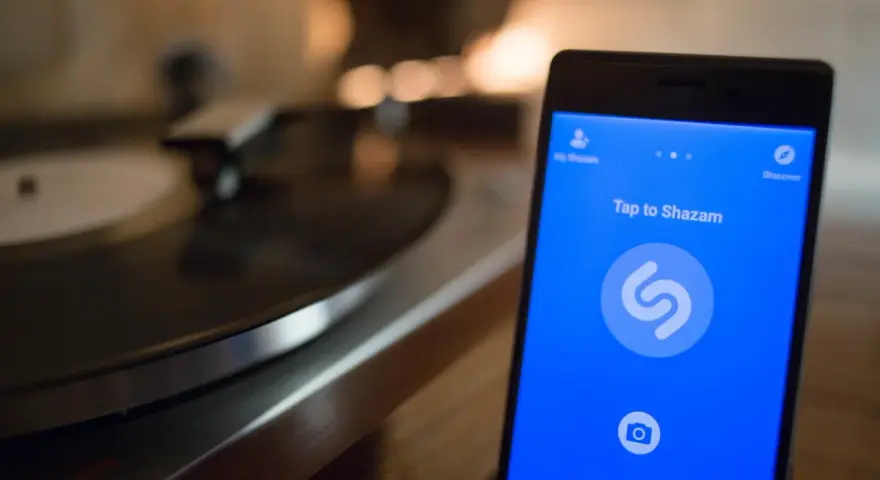 Smartphone con app Shazam aperta, pronto a identificare un brano. Sullo sfondo, un giradischi vintage.