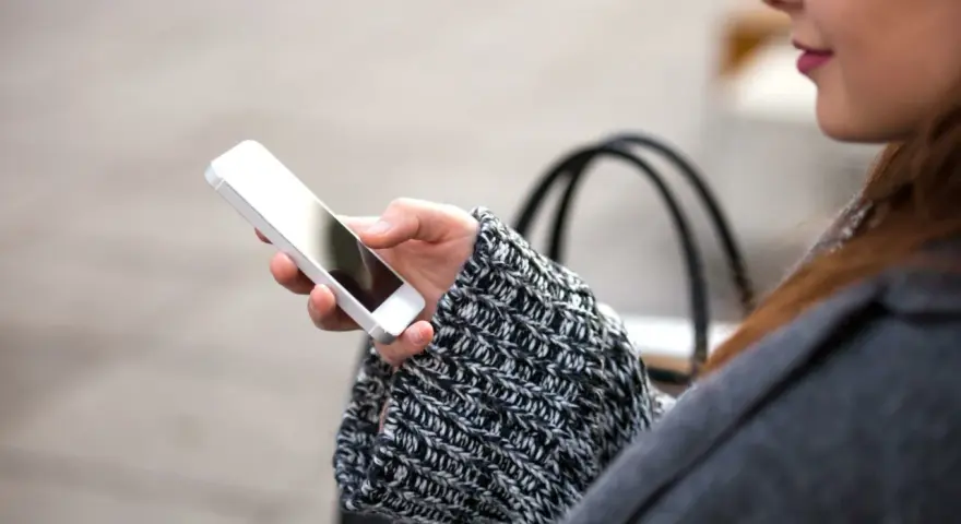 Mano che tiene uno smartphone, con un maglione a trecce e una borsa sullo sfondo. La persona sta utilizzando dati per navigare.