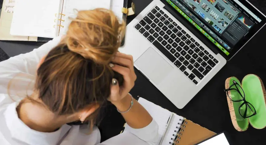 Donna frustrata con la testa tra le mani, laptop aperto, agenda e occhiali. La connessione lenta sta rendendo il lavoro impossibile.