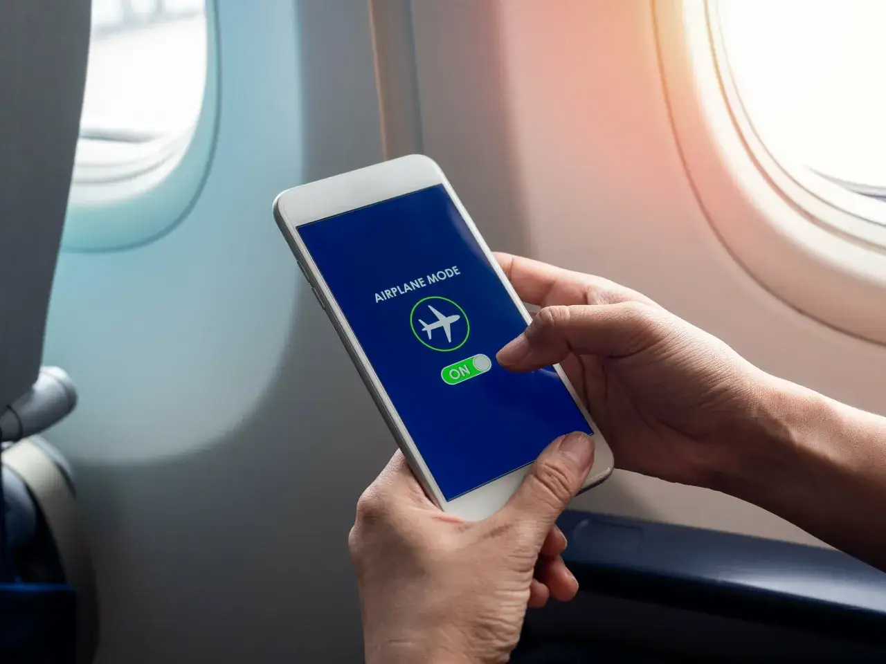 Mani che attivano la modalità aereo sul telefono, seduti vicino all'oblò di un aereo.