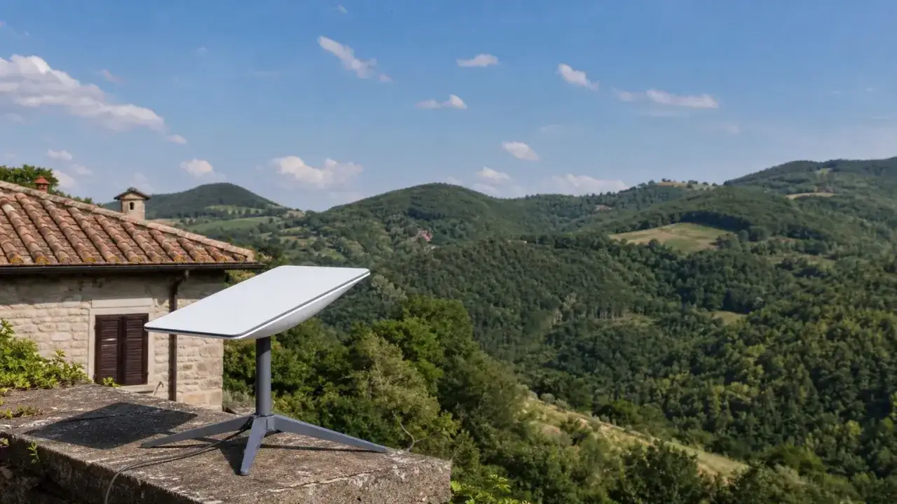 Antenna Starlink Italia su un muro di pietra, con una casa in stile rustico e colline verdi sullo sfondo.