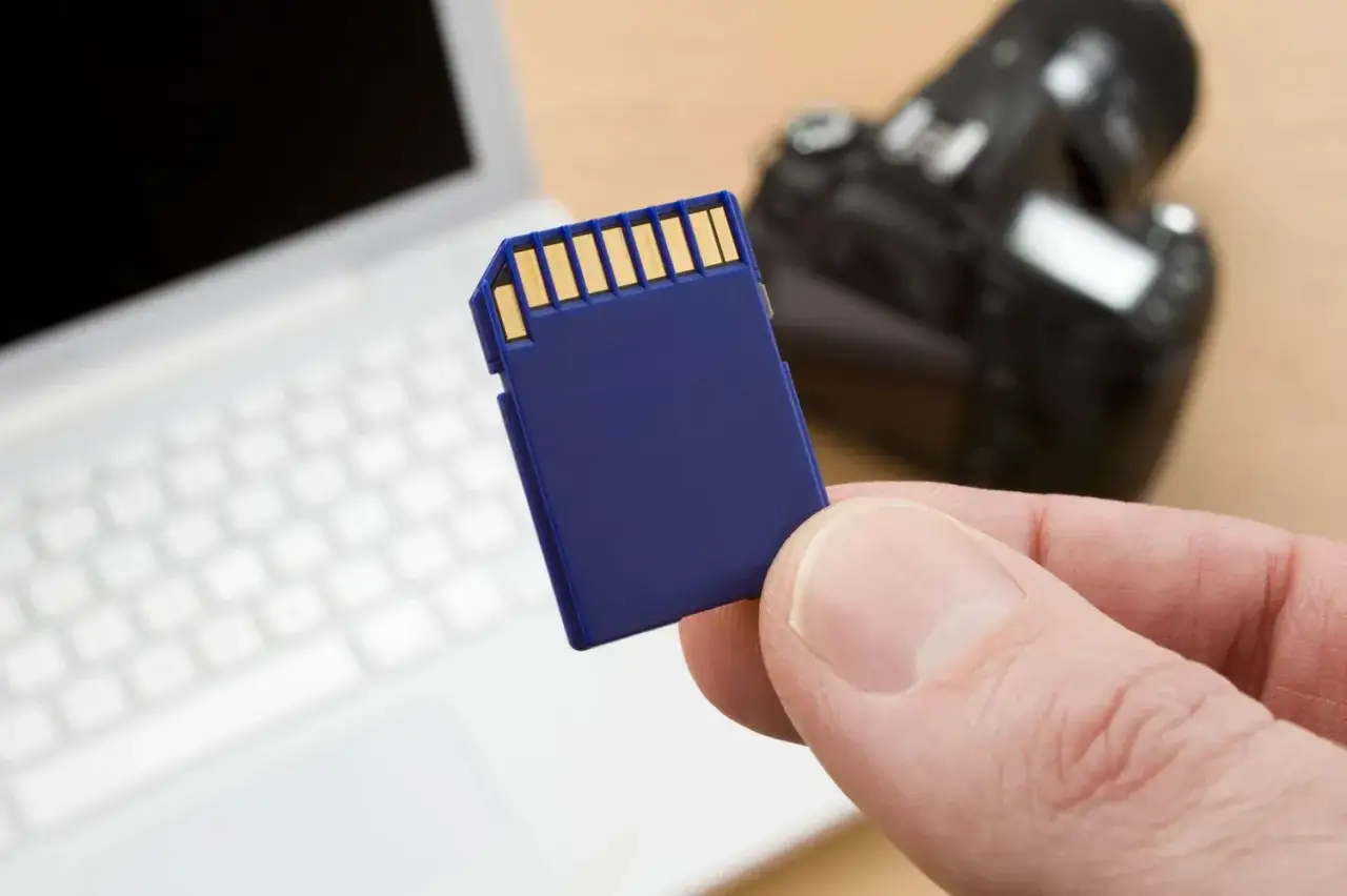 Mano che tiene una scheda SD blu, con un laptop e una fotocamera sullo sfondo. Guida su come formattare scheda SD.