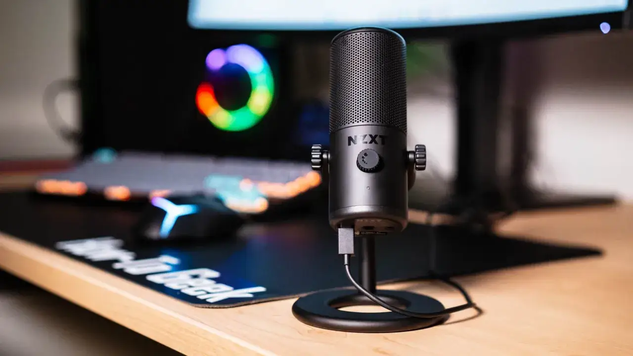 Microfono NZXT pronto per registrare audio PC. Tastiera e mouse RGB sullo sfondo, per un setup da gaming o streaming.
