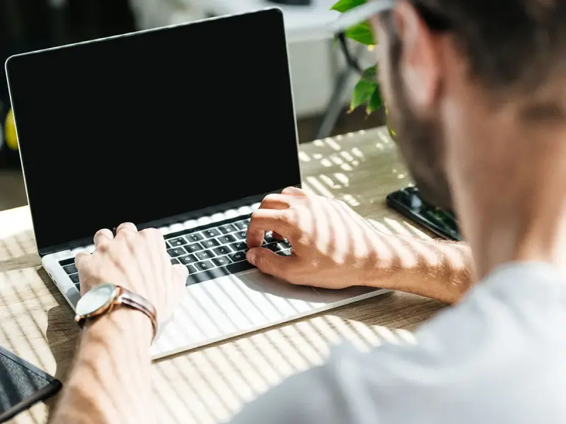 Uomo al lavoro su un laptop, monitora la temperatura CPU. Luce solare filtra attraverso le persiane.