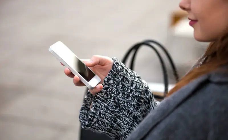 Mano che tiene uno smartphone, con un maglione a trecce e una borsa sullo sfondo. La persona sta utilizzando dati per navigare.